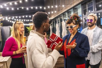 Multi-ethic friends giving a gift box to a woman celebrating birthday in the terrace of a modern