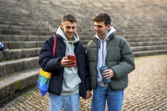 Happy gay couple holding hands, looking at smartphone and enjoying a reusable cup of coffee