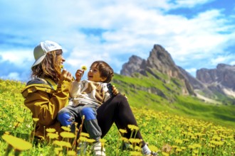 Mother and son playing with dandelions in a field of wildflowers with the seceda mountains in the