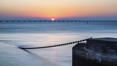 A serene sunset with the sun touching the horizon, beside a calm ocean and a pier extending into