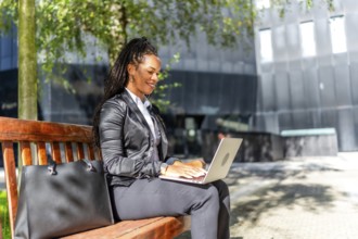 Hispanic elegant businesswoman sitting on city bench working with laptop in the financial district