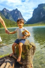 Young boy holding a plush toy and his mother's hand while sitting on a tree stump by lake dobbiaco,