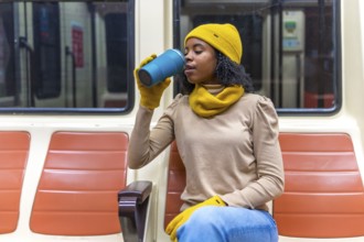 Young woman wearing winter clothes is sitting on a subway train, enjoying a warm beverage from her