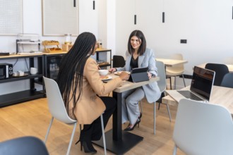 Two businesswomen enjoying breakfast and discussing work using a digital tablet in a modern hotel