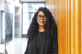 Portrait of a relaxed latin young businesswoman standing and leaning in an office corridor