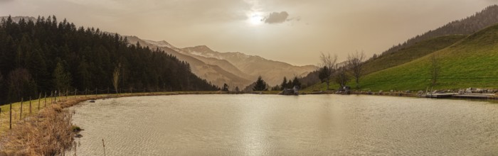 A tranquil lake against a mountain backdrop at sunset with trees and a gentle sky, the small