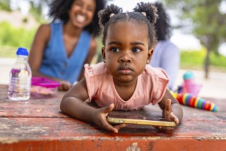 Close-up of a cute African baby girl using mobile phone in the park while her mother and granny