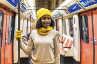 Young black woman wearing yellow wool cap and gloves standing inside subway train, holding handrail