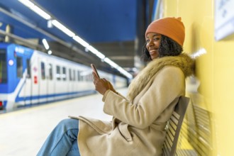 Smiling woman using mobile phone while sitting on bench in subway station with train arriving