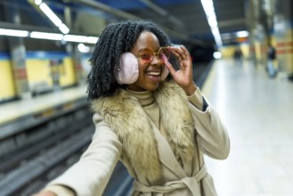 Smiling young woman wearing earmuffs and stylish glasses taking a selfie while waiting for the