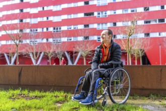 Young man sitting in wheelchair enjoying a sunny day in a modern urban park with a red building in