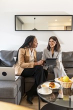 Two businesswomen are sitting on a sofa in a hotel room, using a tablet and laptop during a