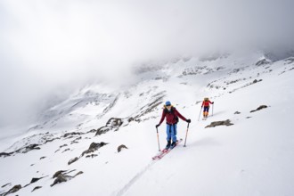 Two ski tourers in a wintry mountain landscape with cloudy mountain peaks, ascent to Piz Laviner,
