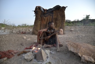 Young shepherdess from the Himba people sitting at a campfire in front of her hut, Ombombo,