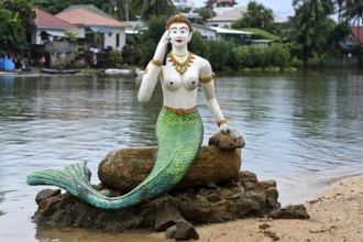 Greeting mermaid statue with white upper body and green tail fin sits on a stone on the shore,