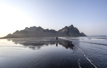 Tourist at black lava beach, sandy beach and sea, mountains Klifatindur reflected in the water,