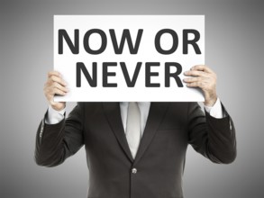 A business man holding a paper in front of his face with the text now or never