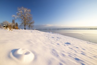 Snow-covered hilly landscape with trees and a frozen body of water under a clear sky, Seegarten,