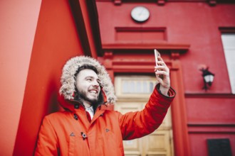 Handsome young male student with smile and beard stands on background red wall, facade of