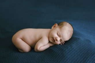 Naked newborn sleeping on a blue blanket with arms and legs tucked in.