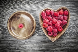 Round and heart-shaped bowls with juicy raspberries on wooden board
