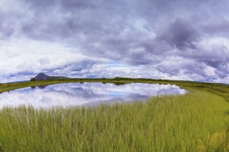 Small lake surrounded by green meadow. In the smooth water of cold lake reflects cloudy sky. Summer