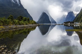 Mirror-smooth water of the Milford Sound fjord reflects mountains. Sunlight breaks through storm