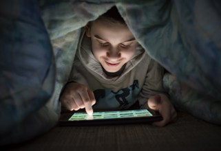 Child watching his tablet in the bed. Illuminated child face from device screen. Smiling Boy under