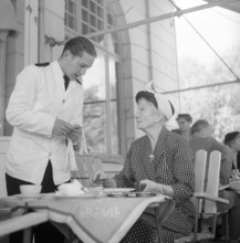 1950s restaurant. An elderly lady is having a conversation with the waiter when settling the bill.