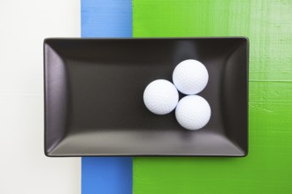 Black ceramic dish with golf balls on over white, blue and green wooden table, rectangle dish