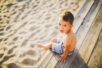 Little Caucasian boy child sitting on wooden pier sandy beach, summer time, sea holiday near water.
