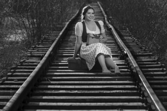 Woman in traditional dirndls sits on railway tracks, relaxed and smiling, black and white picture,