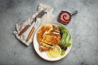 Crispy panko breaded fried chicken fillet with green salad and lemon cut on plate on gray rustic