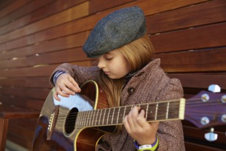 Blond kid girl playing guitar with winter beret and coat on wooden background