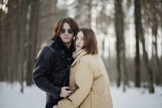 Couple poses in a snowy forest, looking at camera, dressed in winter jackets and scarves