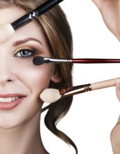 Many hands with cosmetics brushes applying make-up to glamour woman, over white background