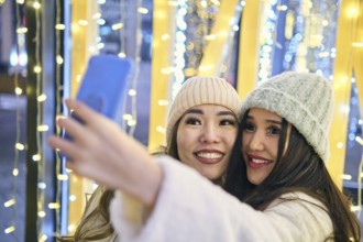 Two girlfriends asian girls in white coats are having fun hugging and selfie on mobile phone, new