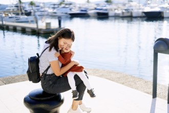 Young woman is hugging her daughter tight while sitting on a pier in a marina.