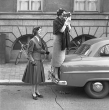 In the 1950s. An elderly woman with a film camera has stood on the bumper of a car to get a better