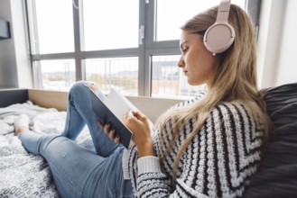 Blonde young caucasian woman lying on her dorm room bed next to a window putting on headphones,