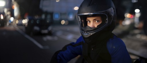 A person in a helmet and motorcycle gear standing by the street with urban lights in the background