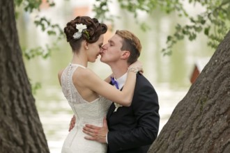 Just married couple kissing between tree trunks and pond