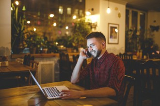 Young handsome Caucasian man with beard and toothy smile in red shirt works behind laptop, hands on