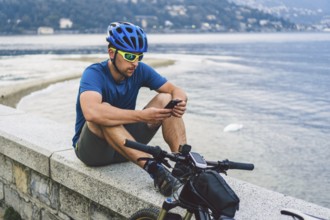 The theme of tourism and travel in Italy. A male cyclist uses a phone on the shore of Lake Como.