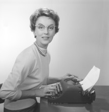 Office girl in the 1950s. A young girl at her typewriter is wearing typical fifties glasses and