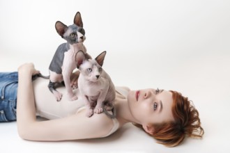 Two friendly Sphynx Hairless Cat, sitting on redhead young woman lying on back on white background.