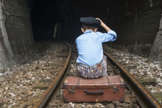 Child in vintage clothes sits on railway road in front of a tunnel