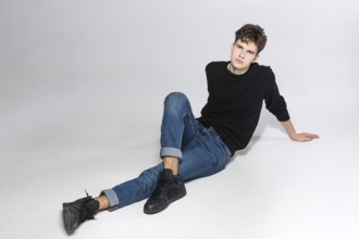 Stylish guy posing in studio sitting on the floor in denim pants and black sweatshirt. Male student