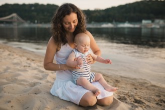 Young mother sitting on the beach with one year old baby son. Boy hugging, smiling, laughing,