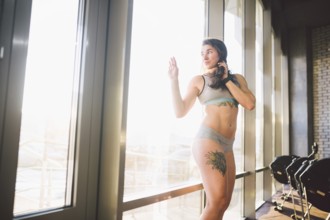 Young caucasian sporty woman with curly long hair poses in sportswear near a panorman window in a